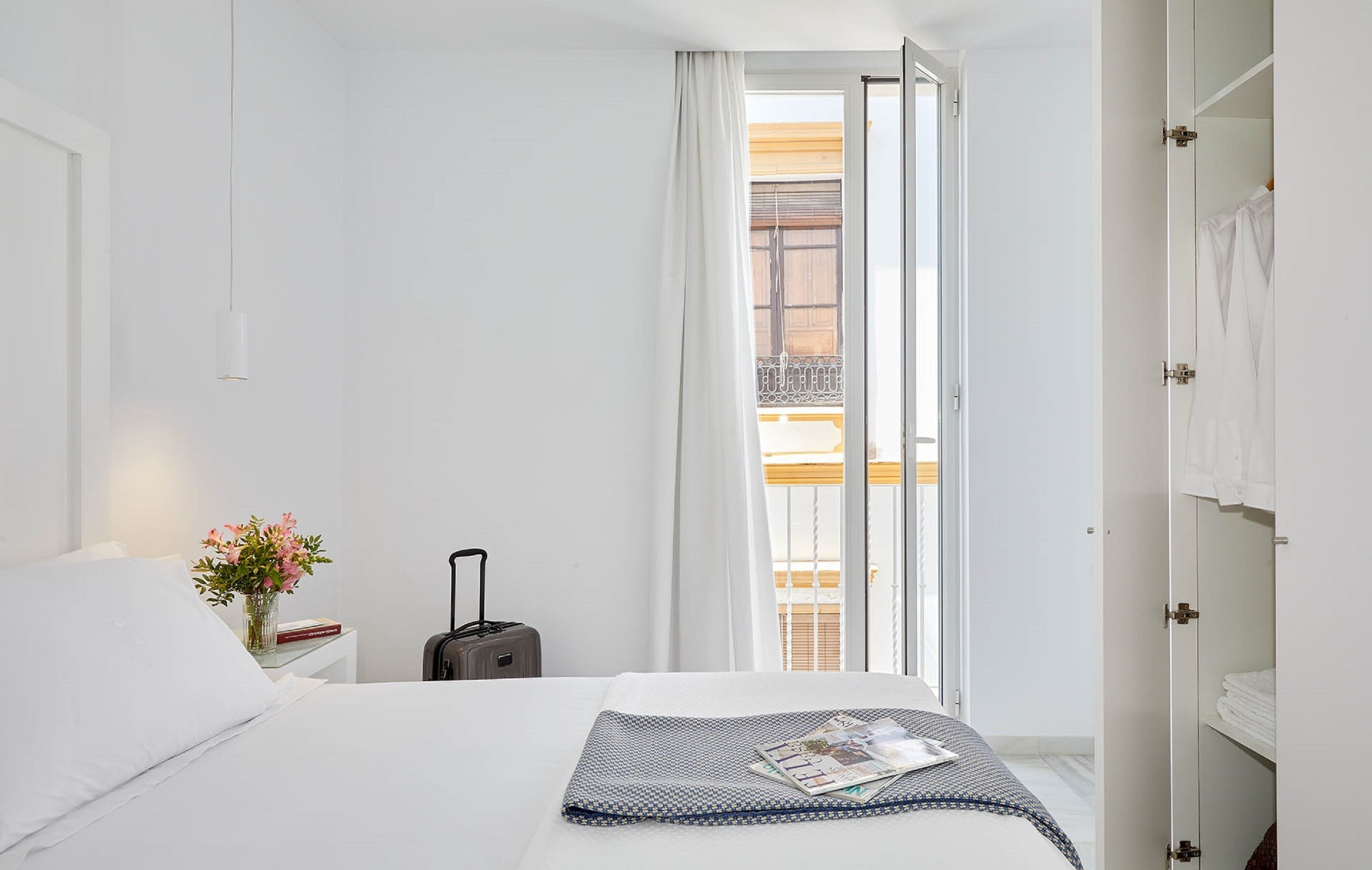 Une chambre d’hôtel avec un lit, une valise, une table de chevet ornée d’un vase de fleurs et une vue sur un balcon.