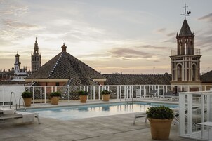A rooftop pool with lounge chairs, potted plants, and a view of historic buildings in the background.