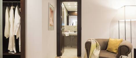A hotel room with a wardrobe, a sofa with a throw blanket, a coffee table with a cup and a bowl of cookies, and a bathroom visible through an open door.