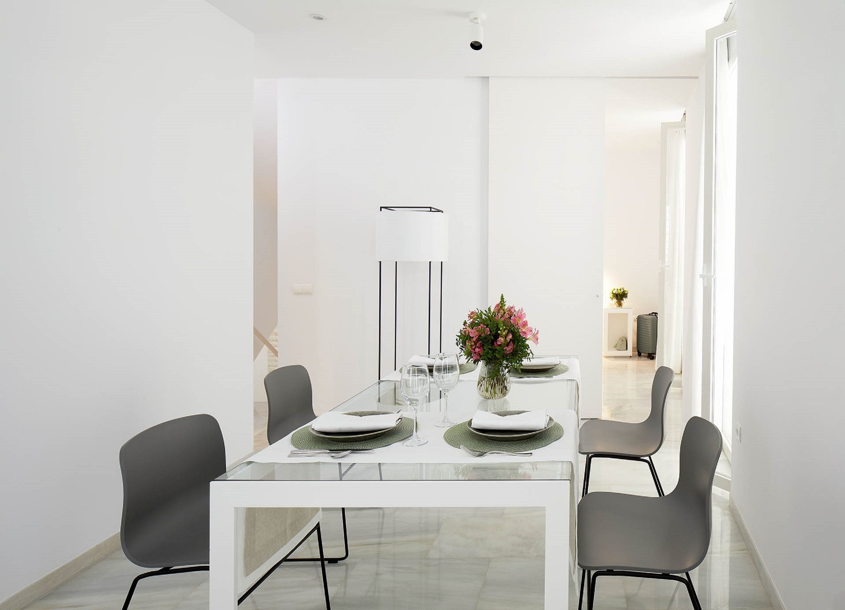 A modern dining area with a glass table, grey chairs, white plates, and a vase of flowers.