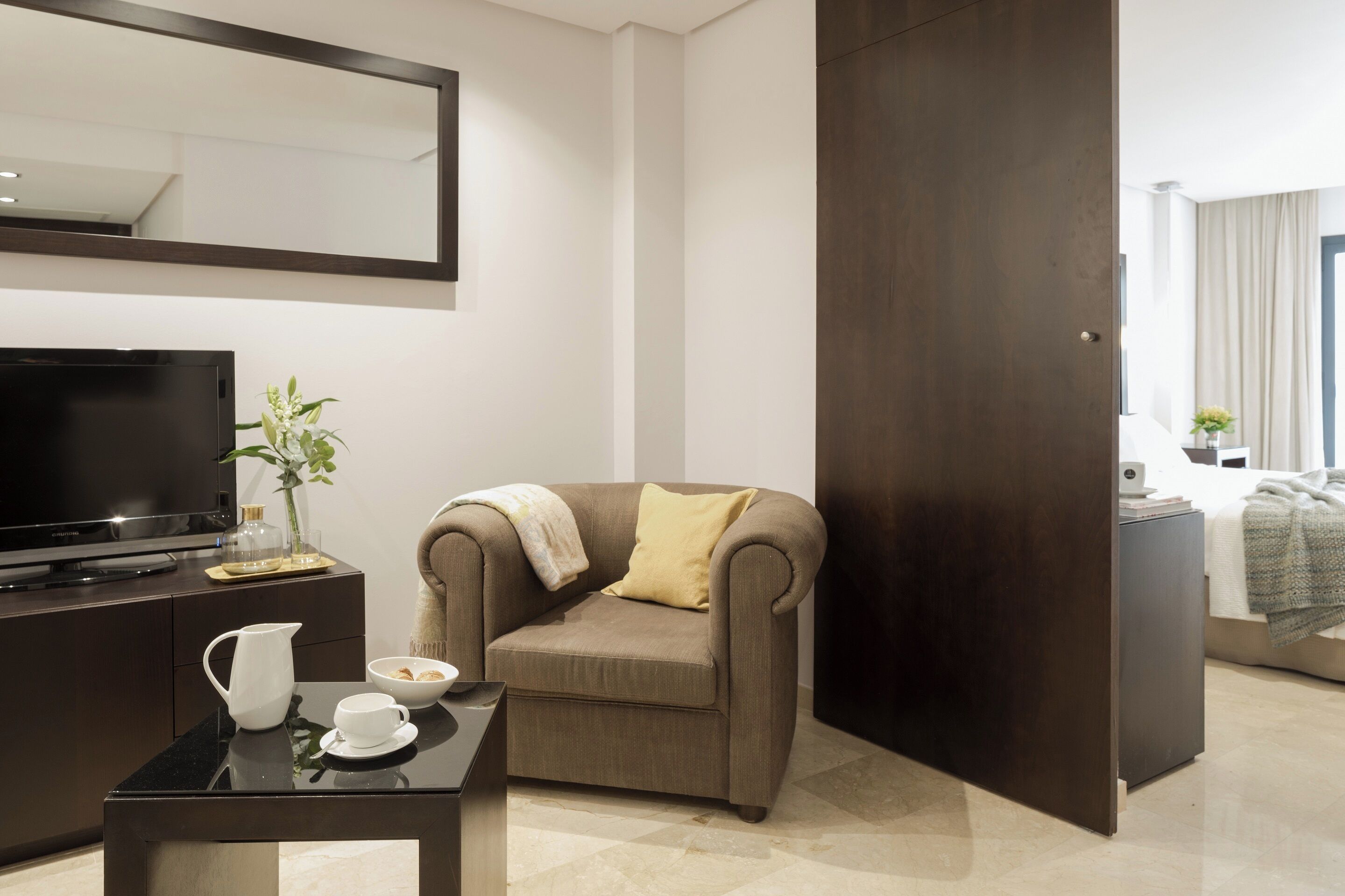 Una habitación de hotel moderna con una televisión de pantalla plana, un sillón cómodo, una mesita de café y un espejo grande.