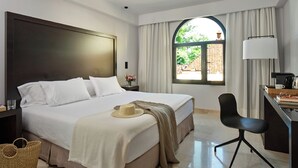 A modern hotel room with a large bed, a desk with a lamp, a chair, and a view of greenery through a window. - Hotel Fernando III (Seville)