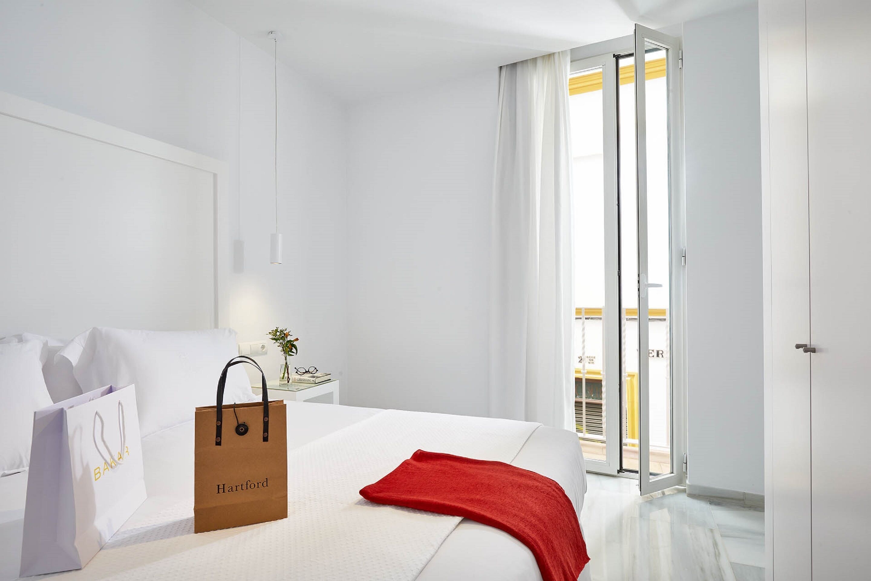 A hotel room with a bed, a red throw blanket, a white pillow, a white wall, a nightstand with a lamp, and a sliding glass door to the balcony.