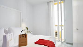 Habitación de hotel con una cama, un plaid rojo, una almohada blanca, una pared blanca, una mesita de noche con lámpara y una puerta corrediza de vidrio al balcón.