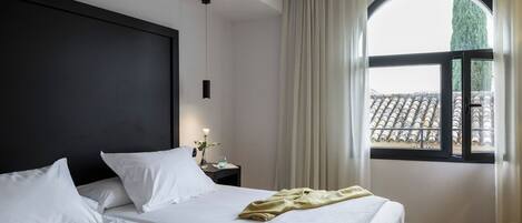 Habitación de hotel con dos camas, un ventanal con cortinas, una mesita de noche con lámpara y vista a un tejado de baldosas.