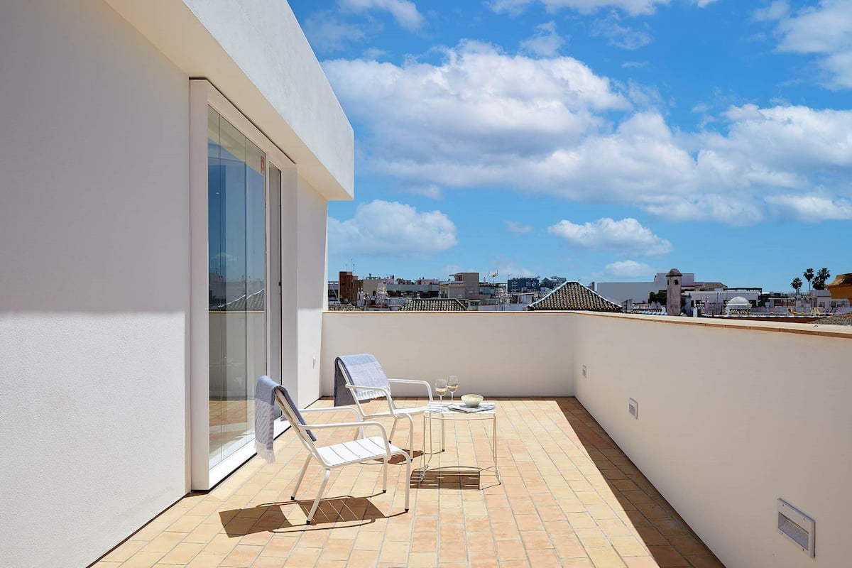 A rooftop terrace with two white lounge chairs, a small table with a bowl and glasses, and a glass door leading to an indoor area.