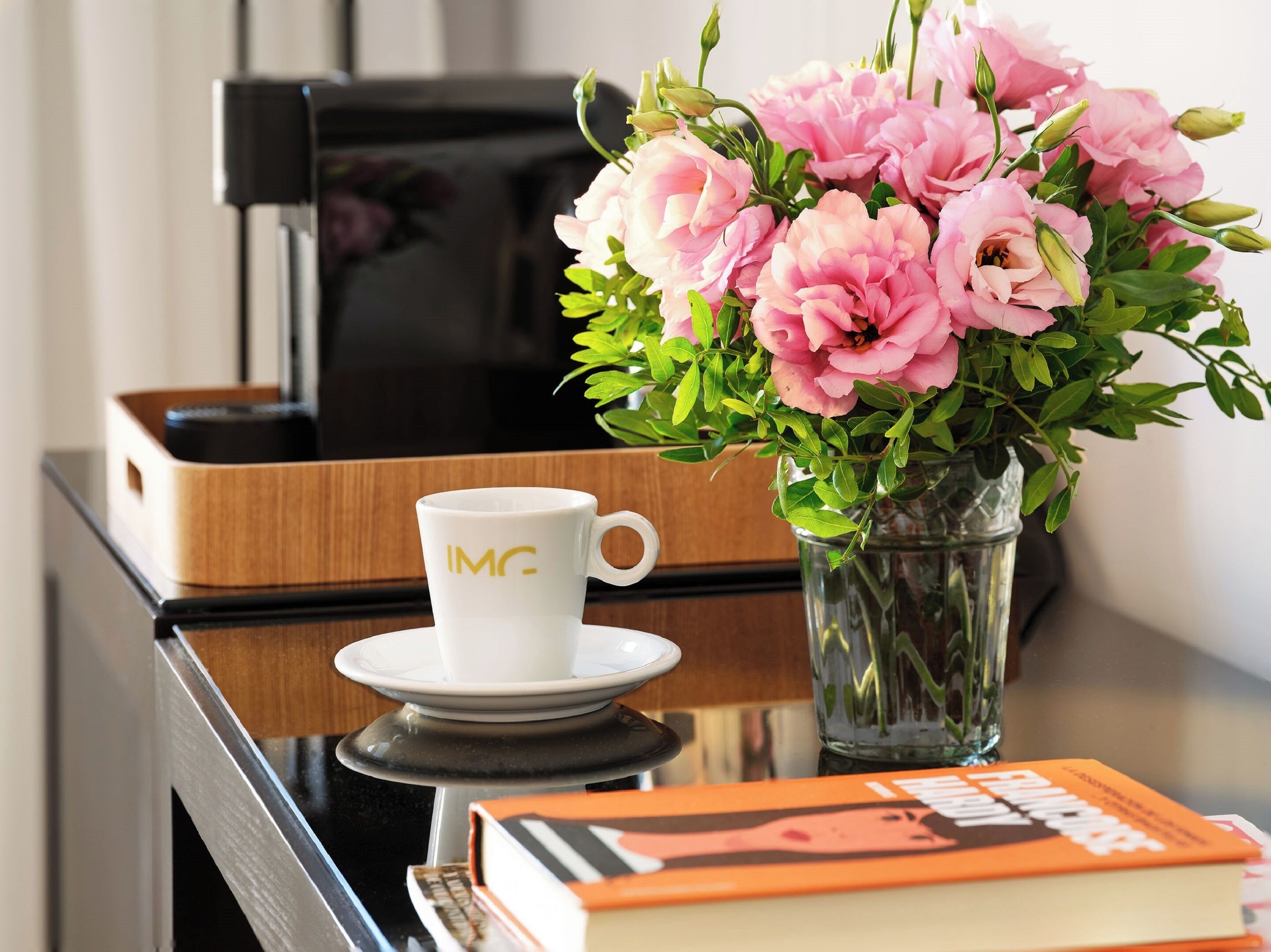 Una tazza da caffè con il logo "IMG", un libro e un vaso con fiori rosa su un tavolo.