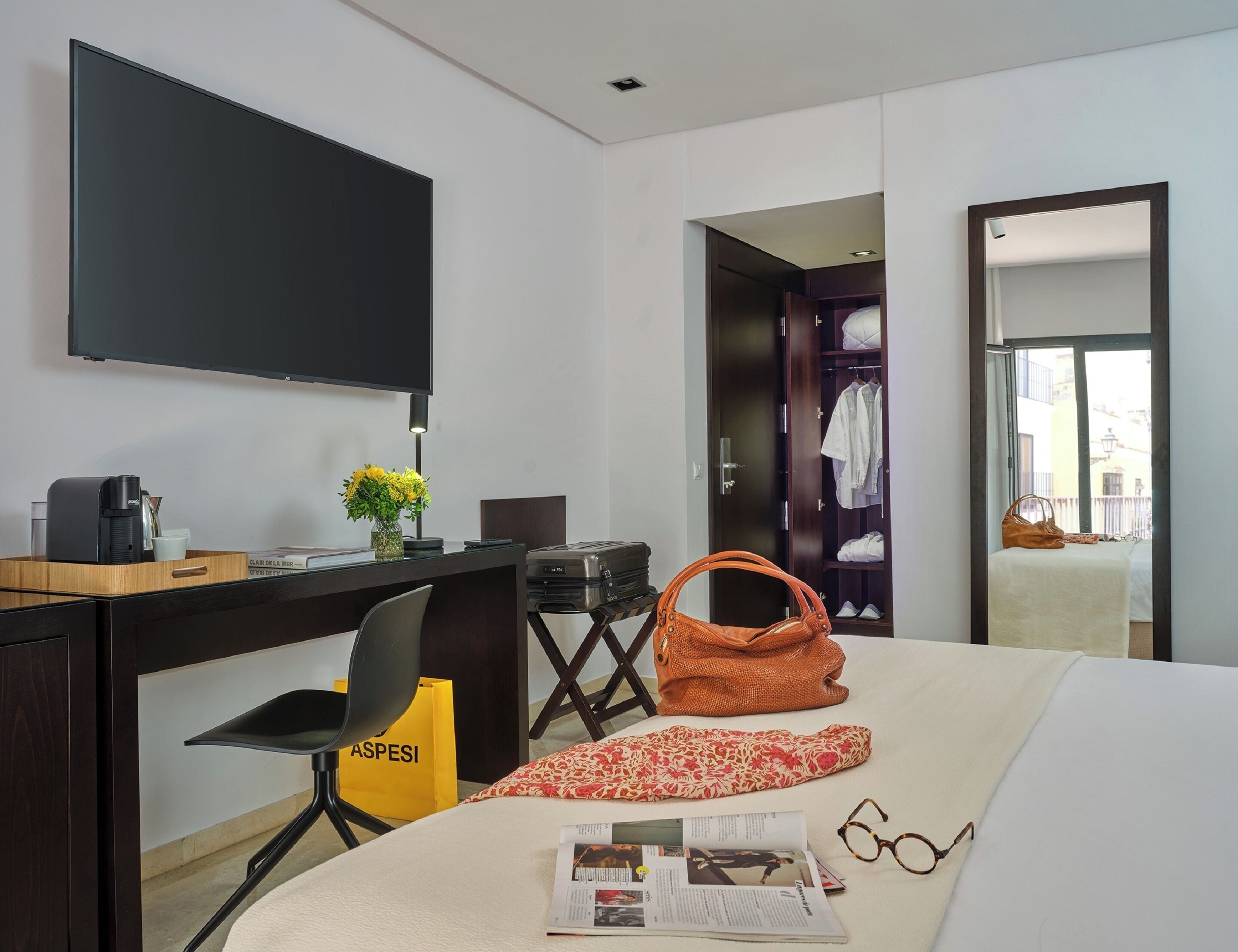 Habitación de hotel con cama, escritorio con cafetera, televisor, armario y bolso sobre la cama.