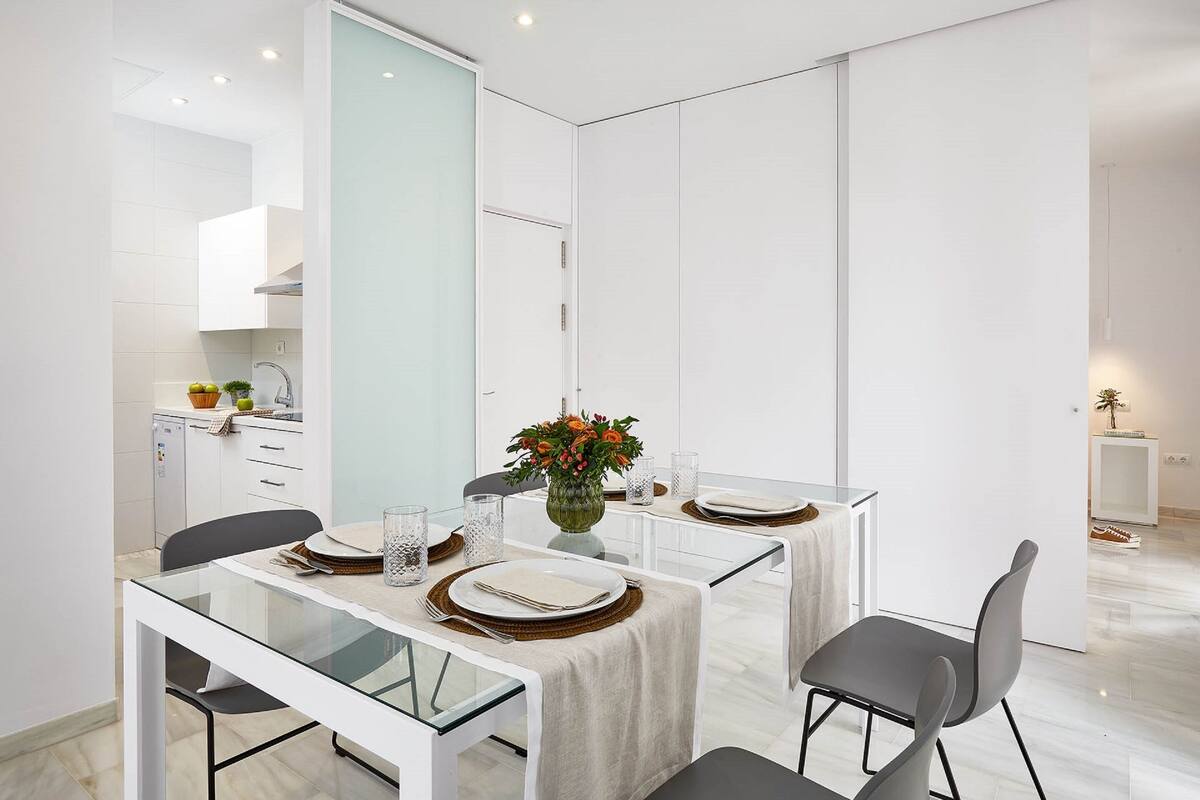 A modern dining area with a glass table, white walls, and a kitchen in the background.