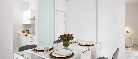A modern dining area with a glass table, white walls, and a kitchen in the background.