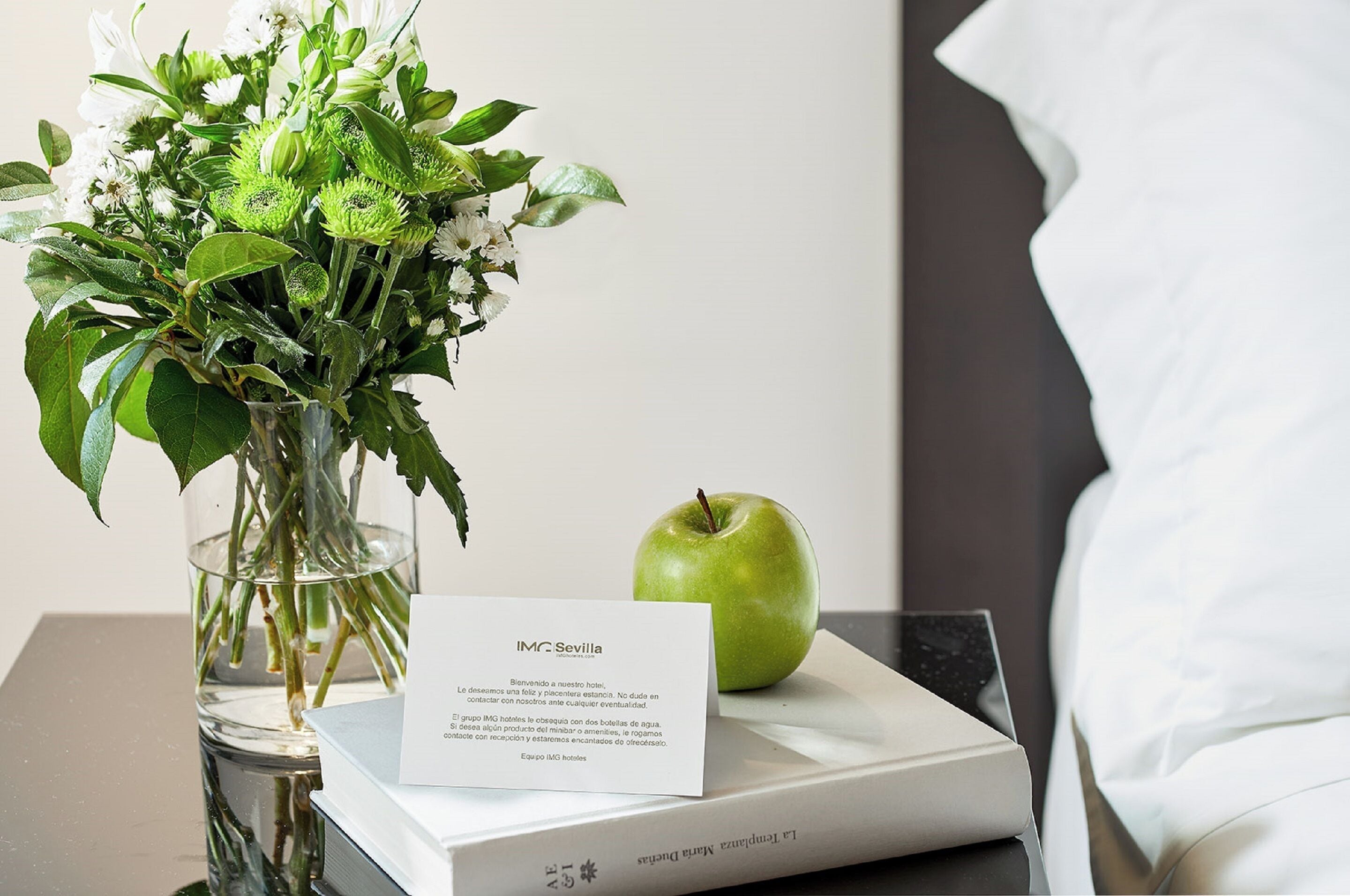 A neatly arranged hotel room with a vase of flowers, a green apple, and a welcome card on a bedside table.