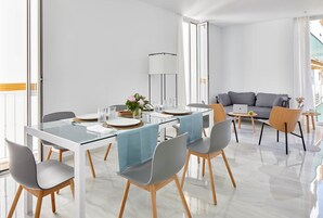 Un comedor moderno con una mesa de vidrio, sillas grises y mantel azul. Se observa un sofá y una lámpara al fondo.