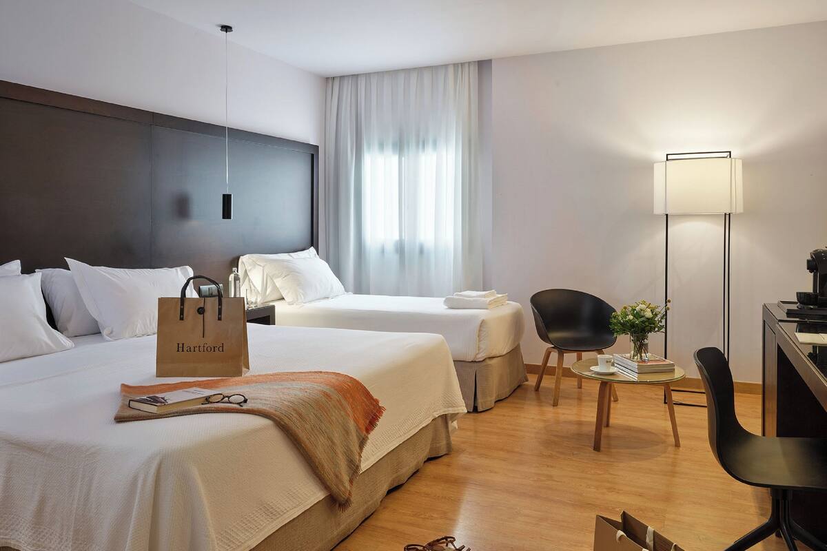 A hotel room with two beds, a desk, a chair, a lamp, and a bag on the floor.
