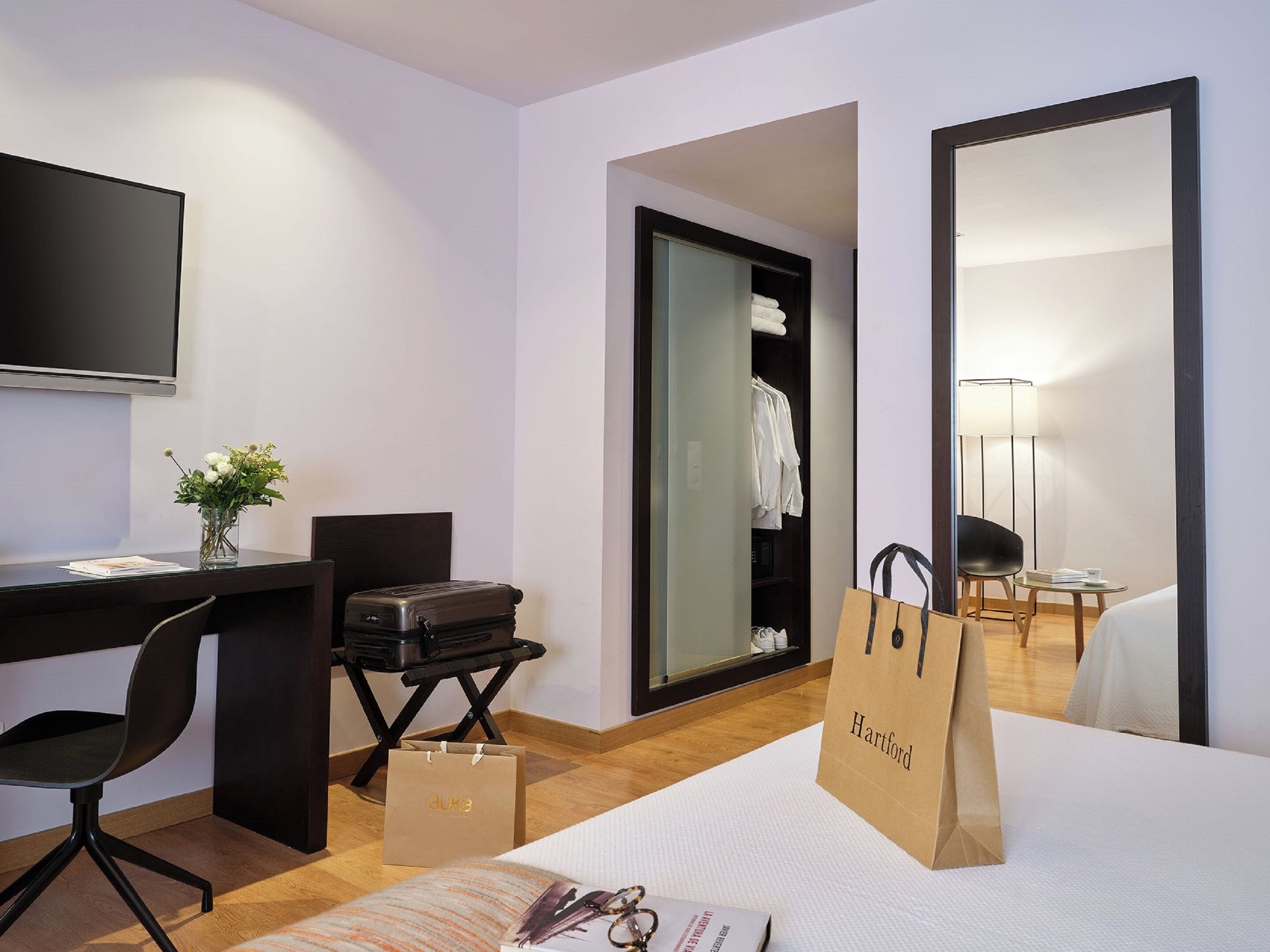 A hotel room with a desk, chair, bed, wardrobe, and a mirror. There are two shopping bags on the bed.