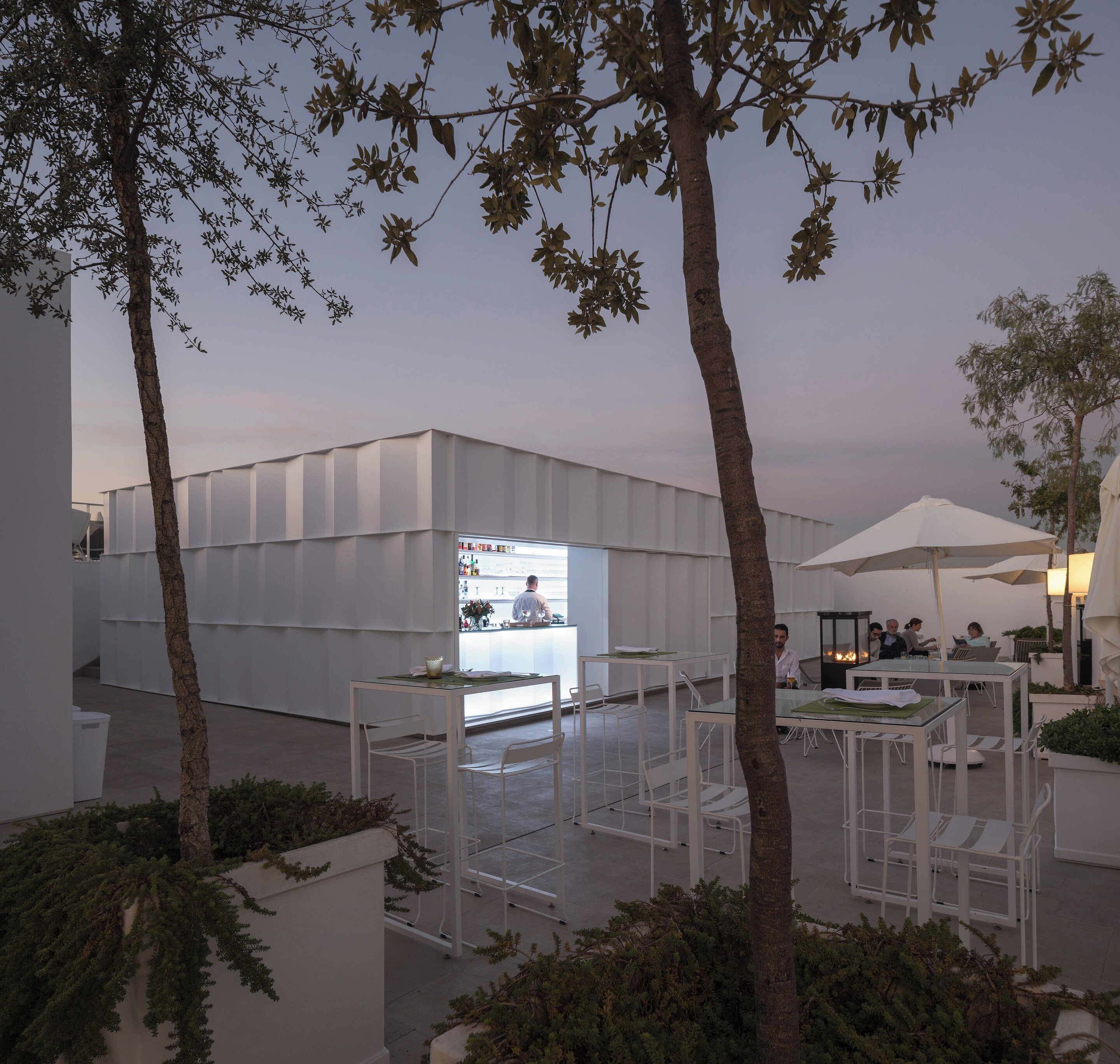 An outdoor dining area with white tables and chairs, surrounded by trees and a modern building with a glass facade.