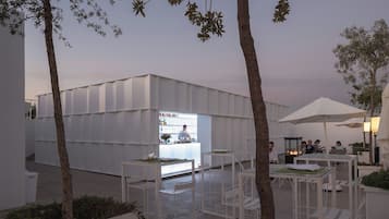 Un área de comedor al aire libre con mesas y sillas blancas, rodeada de árboles y un edificio moderno con fachada de vidrio.