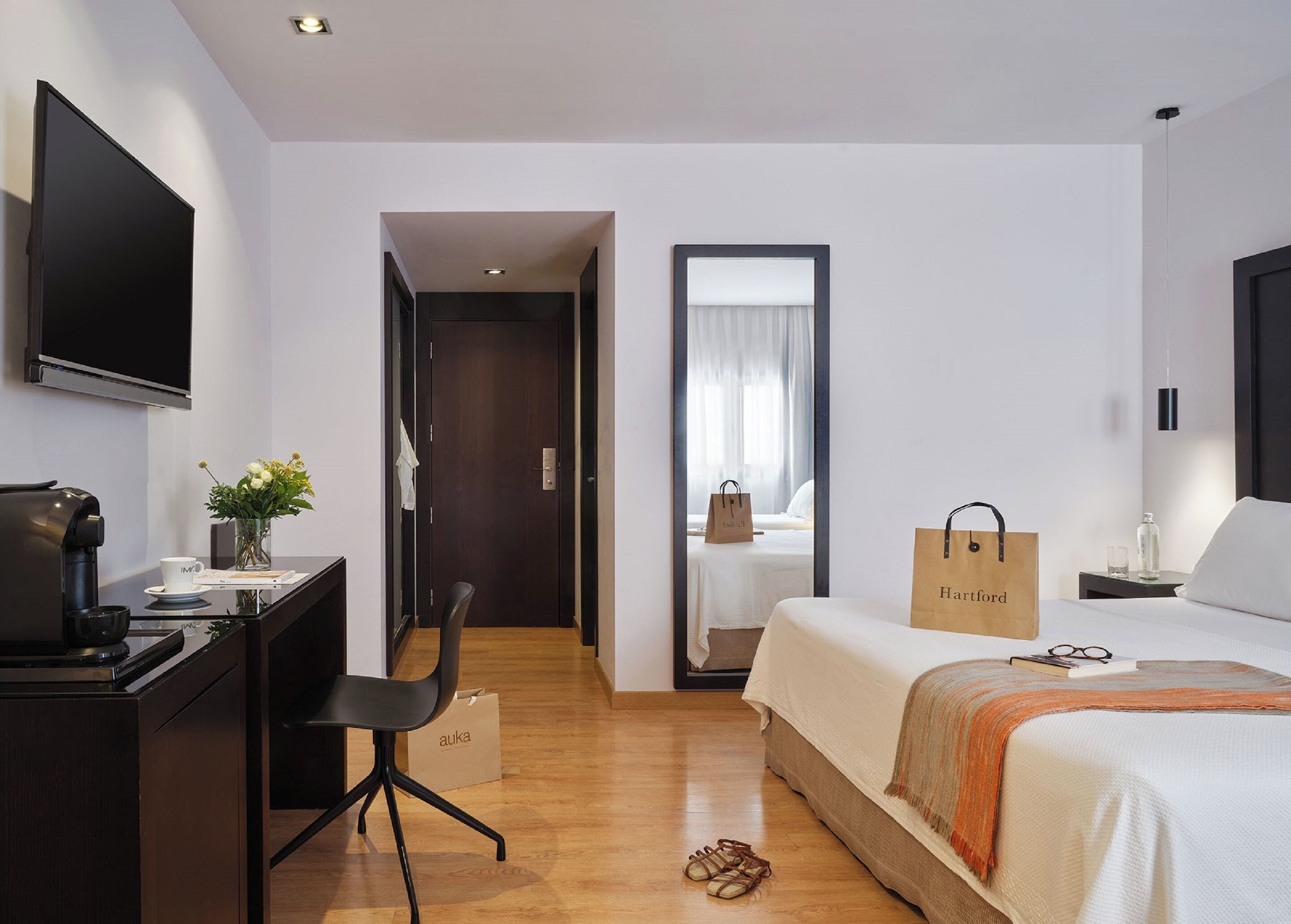 A hotel room with a bed, a desk with a coffee maker, a chair, a TV, and a window with curtains.