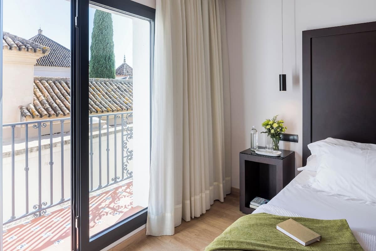A hotel room with a bed, a nightstand with a vase of flowers, a window with curtains, and a view of buildings outside.