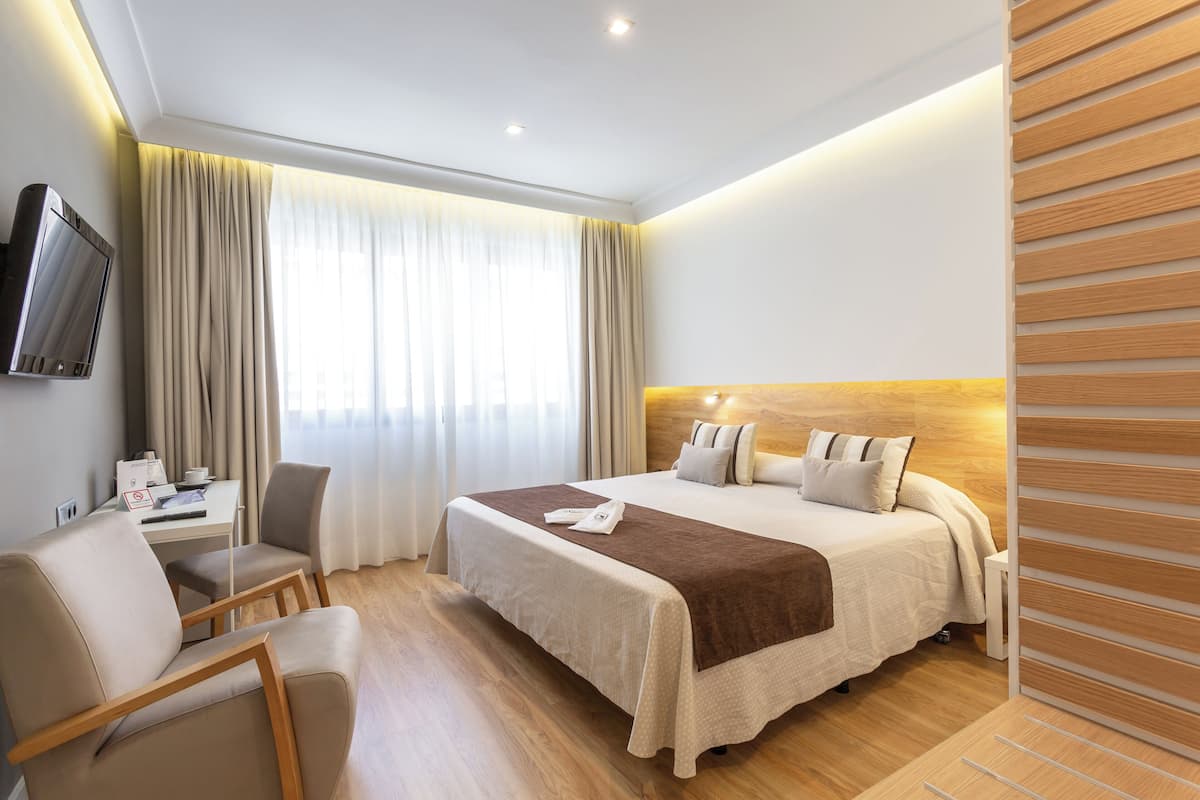 Habitación de hotel con una cama, un escritorio con silla, un televisor montado en la pared y una ventana con cortinas.