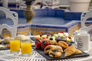 Una mesa con una gran variedad de pasteles, frutas y zumos, con vistas a una fuente.