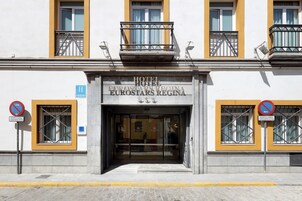 La entrada del Hotel Eurostars Regina con fachada de vidrio transparente, un balcón con barandilla de metal y un letrero que indica que no hay estacionamiento excepto para carga y descarga.