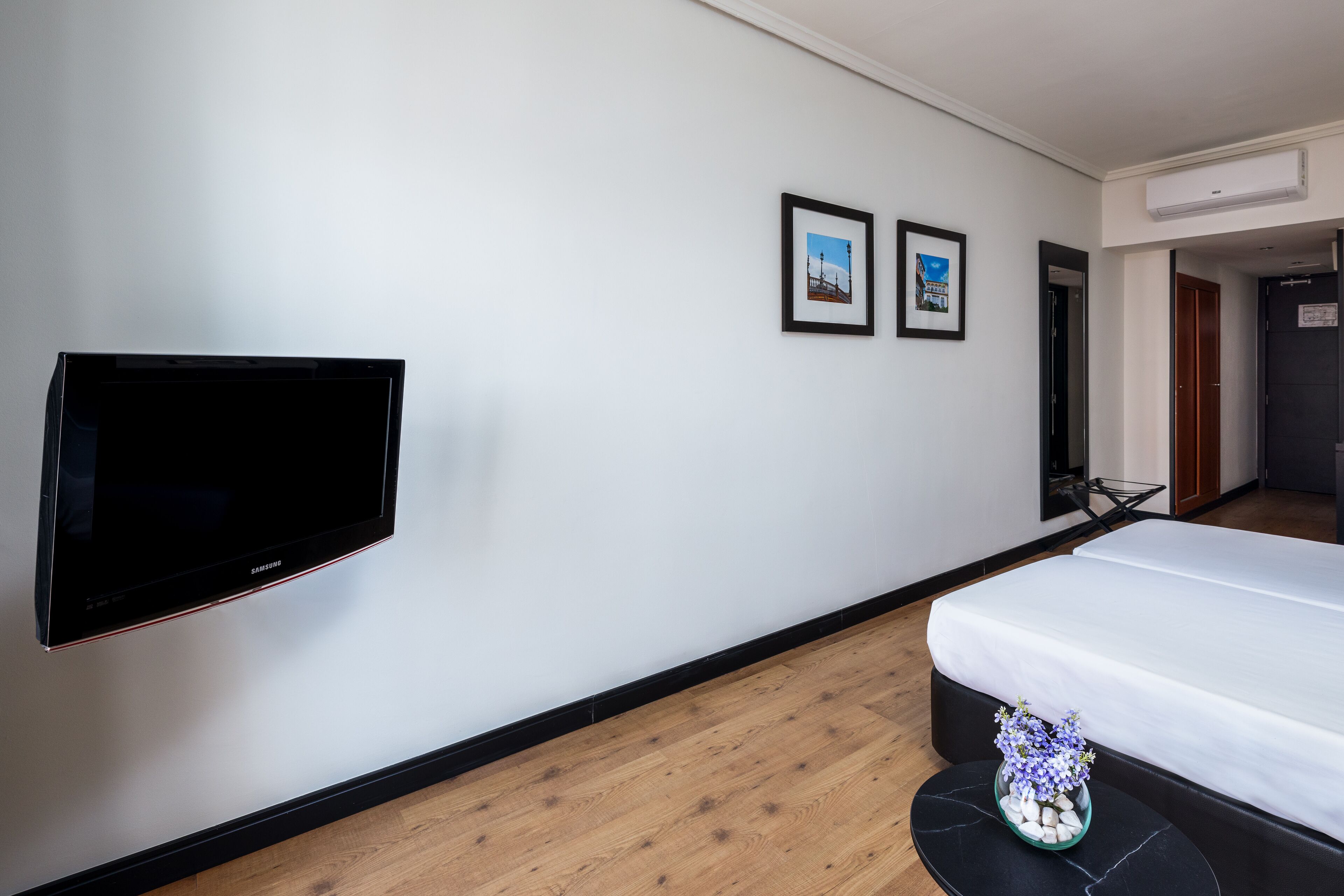 Een hotelkamer met een flatscreen televisie, een bed, een kleine tafel met een bloevas en twee ingelijste foto’s aan de muur.