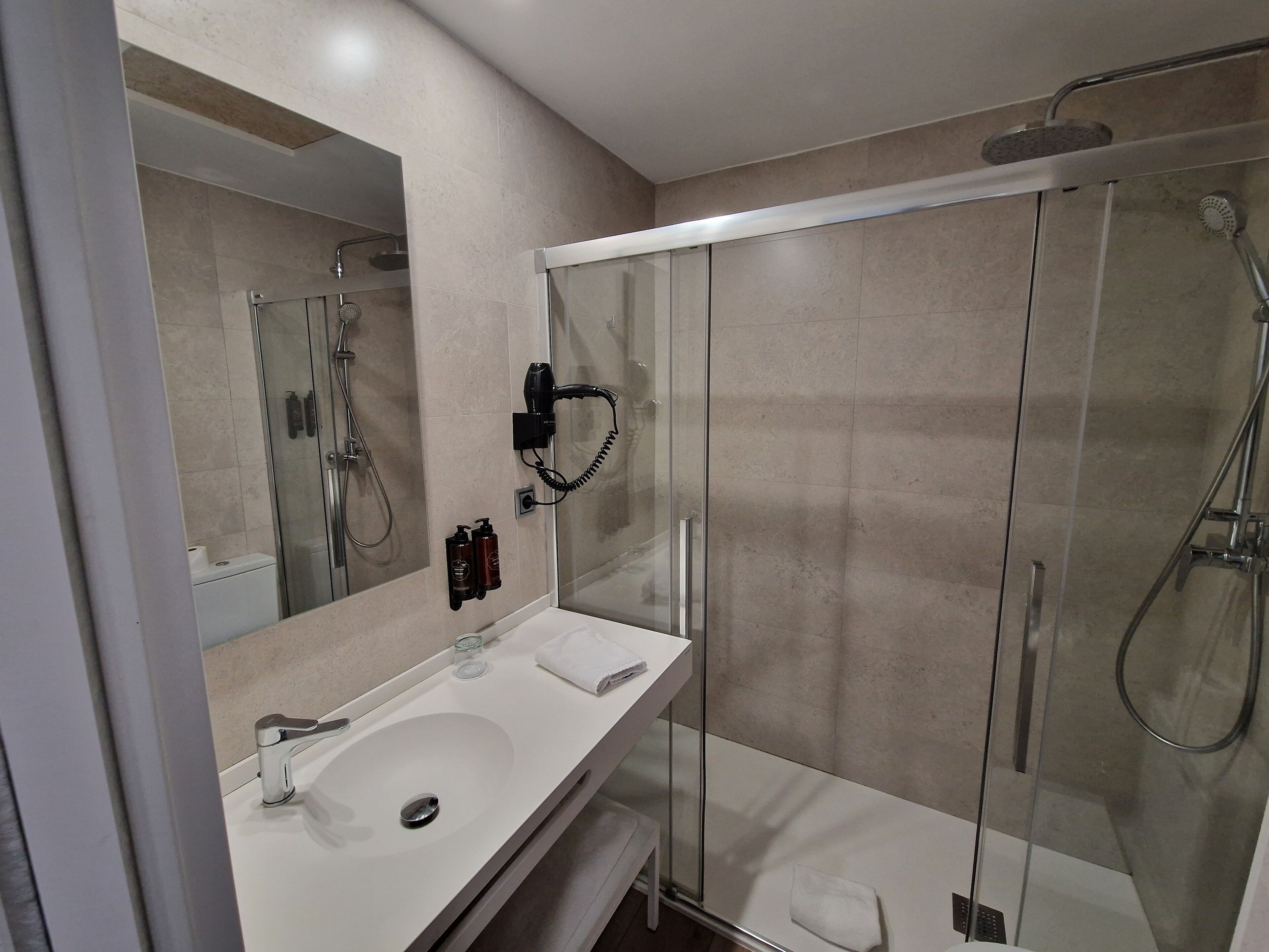 Un baño moderno con cabina de ducha de vidrio, un lavabo blanco y un secador de pelo de pared.