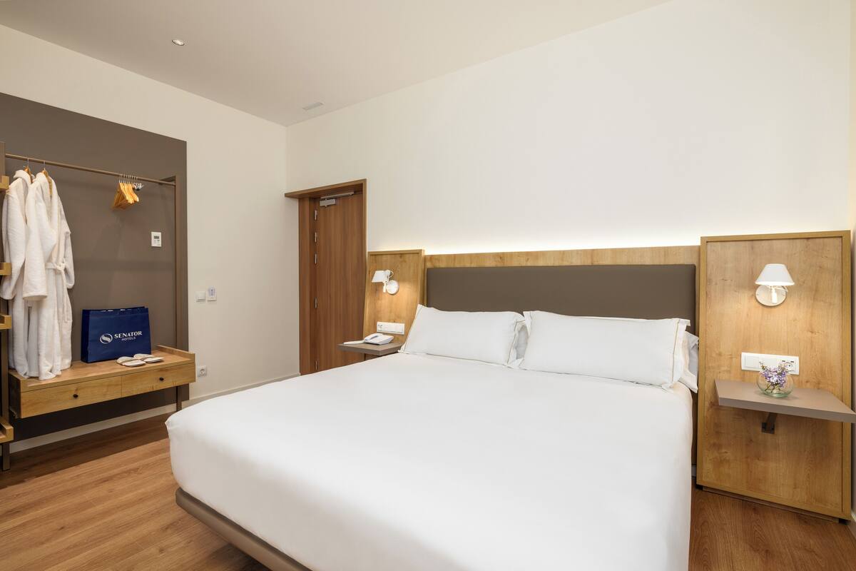A hotel room with a large bed, wooden headboard, bedside tables with lamps, a wardrobe, and a blue bag on the nightstand.