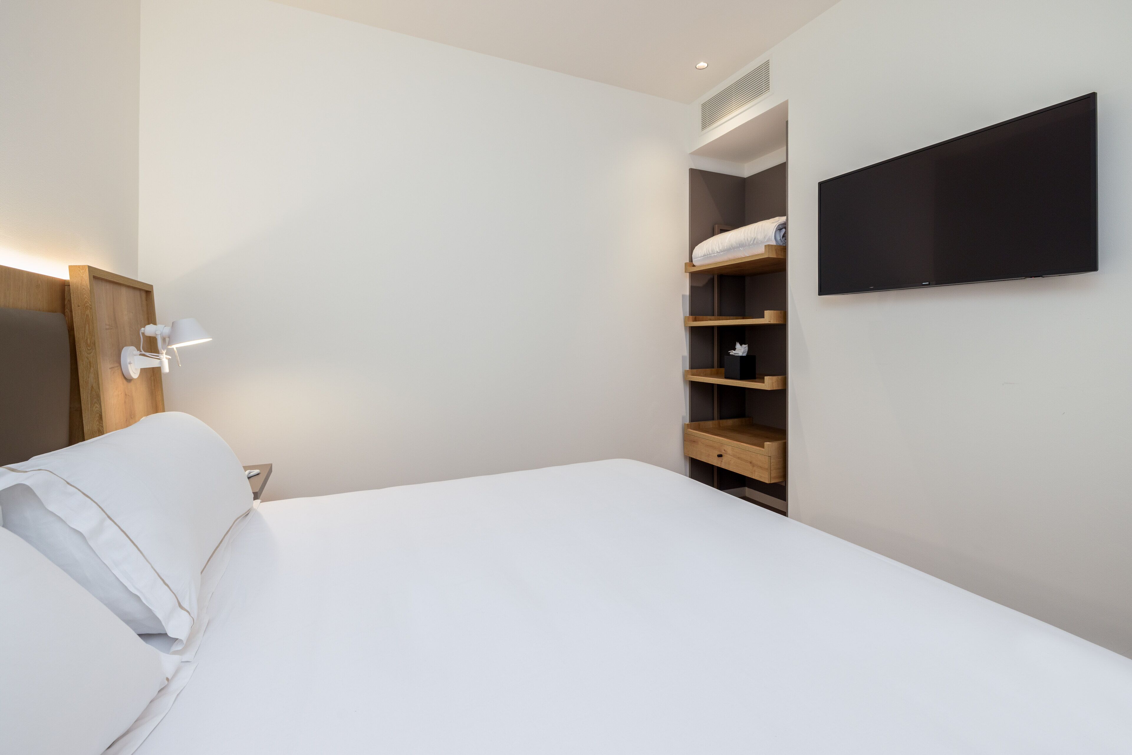 Ein modernes Hotelzimmer mit einem großen Bett, einem an der Wand montierten Fernseher und integrierten Regalen mit Stauraum.