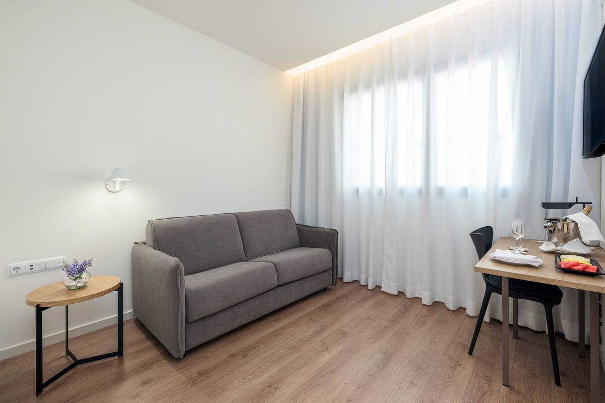 A modern living room with a grey sofa, a small round table with a vase, and a dining area with a table set for two.