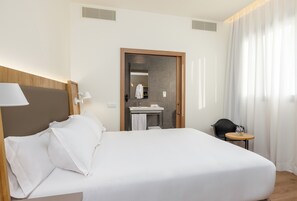 A hotel room with a bed, a bedside table, a lamp, a small round table with a vase, a black chair, a bathroom with a mirror, sink, and towel rack. - Virgen de los Reyes Hotel (Seville)