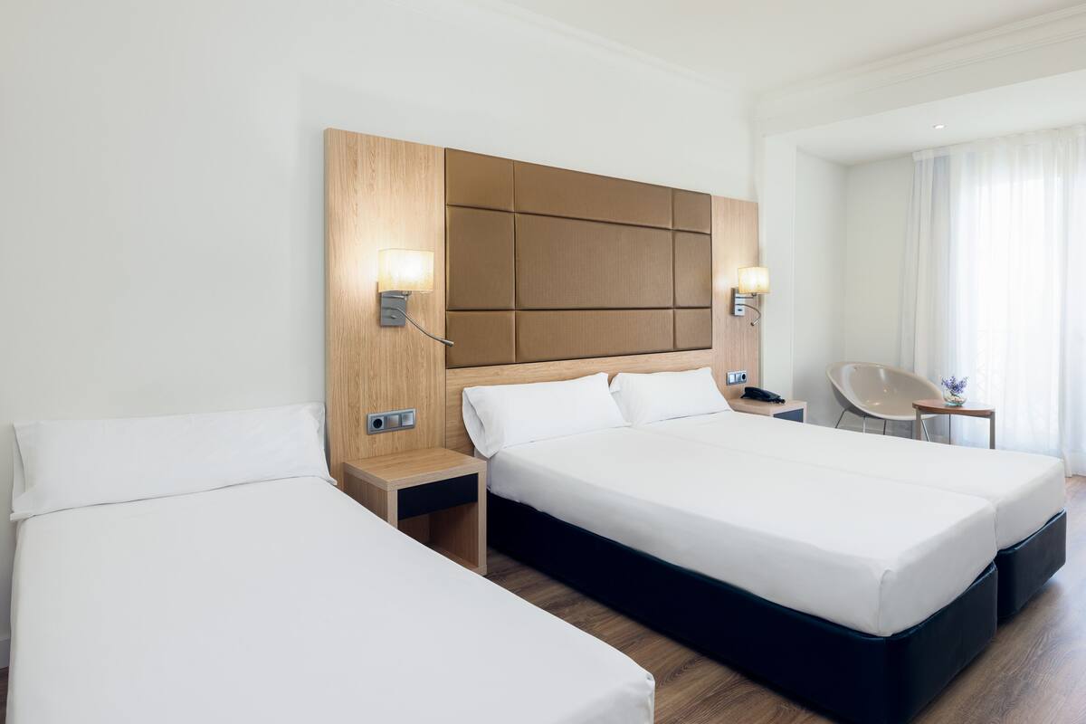 A hotel room with two beds, wooden headboard, bedside lamps, a chair, and a small table.