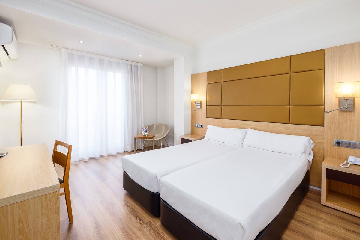 A hotel room with a double bed, a desk with a chair, a small table, and a lamp.