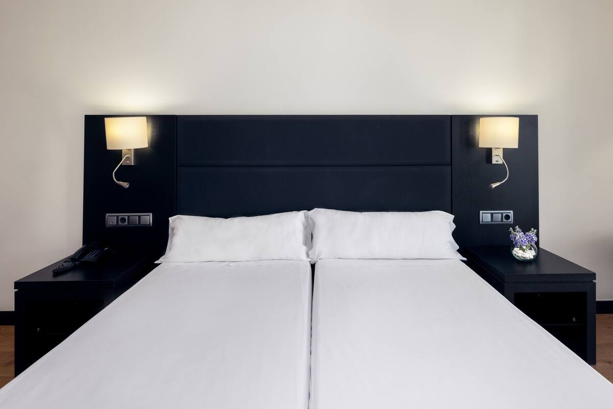 A double bed with white linens, two bedside tables with lamps, and a small vase with flowers on the right table.
