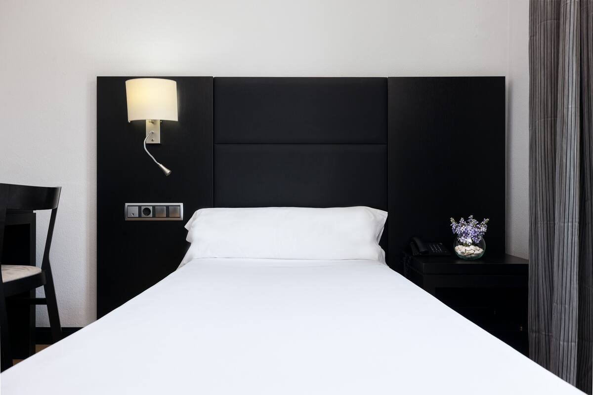A hotel room with a large bed, a bedside table with a lamp, a chair, and a small vase with flowers.