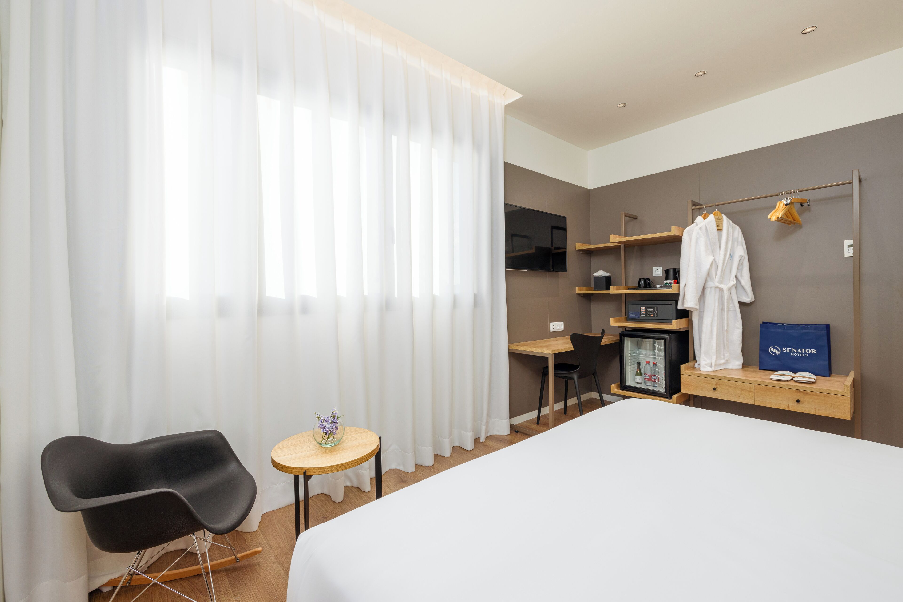 Habitación de hotel con cama, escritorio, silla, una pequeña mesa con un jarrón, minibar, televisión y armario con ropa y bolso.