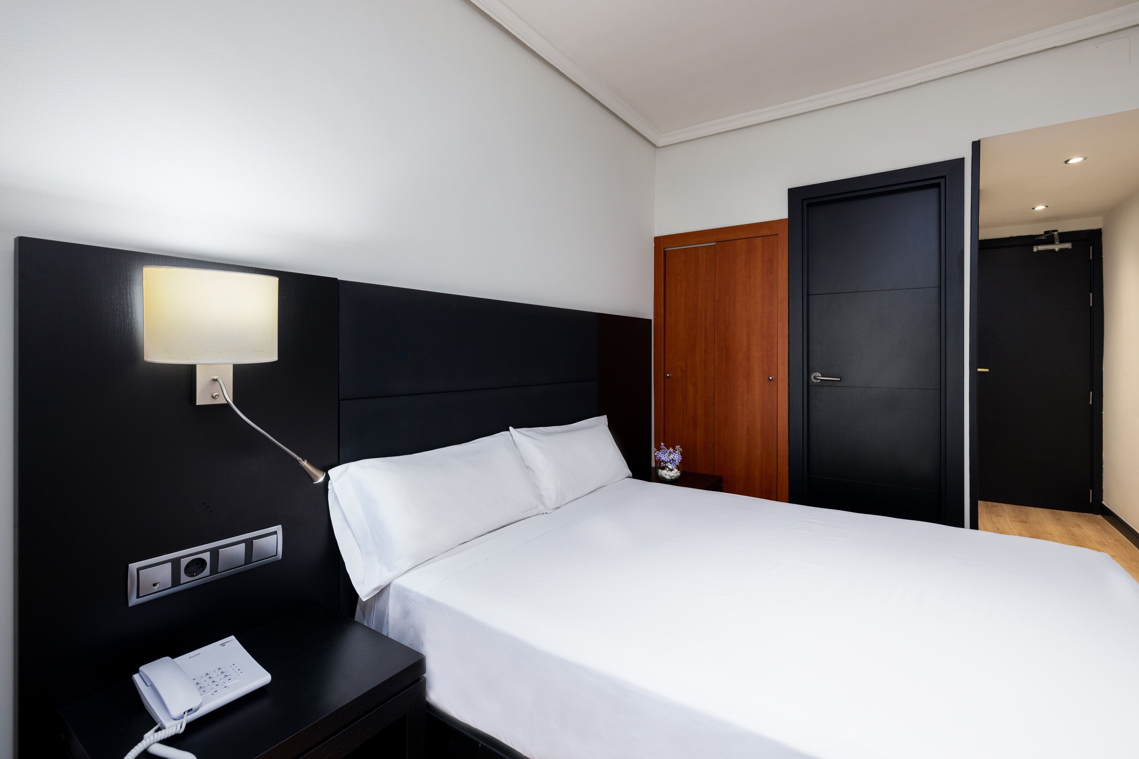 Une chambre d’hôtel moderne avec un grand lit, une table de chevet avec un téléphone, une lampe fixée au mur et une armoire en bois.