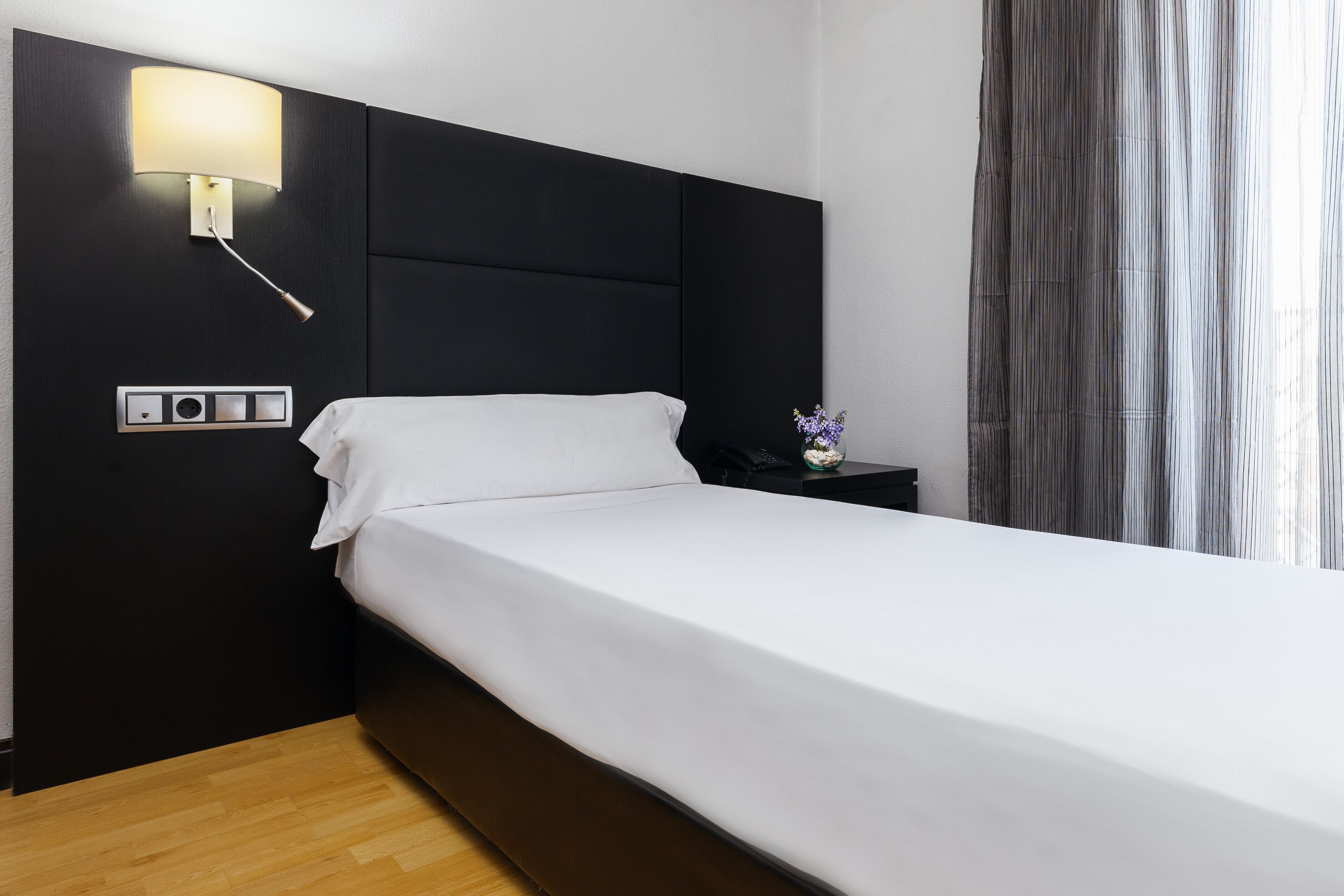 Habitació d'hotel moderna amb un llit gran, una làmpada penjada a la paret, una tauleta de nit i una cortina.