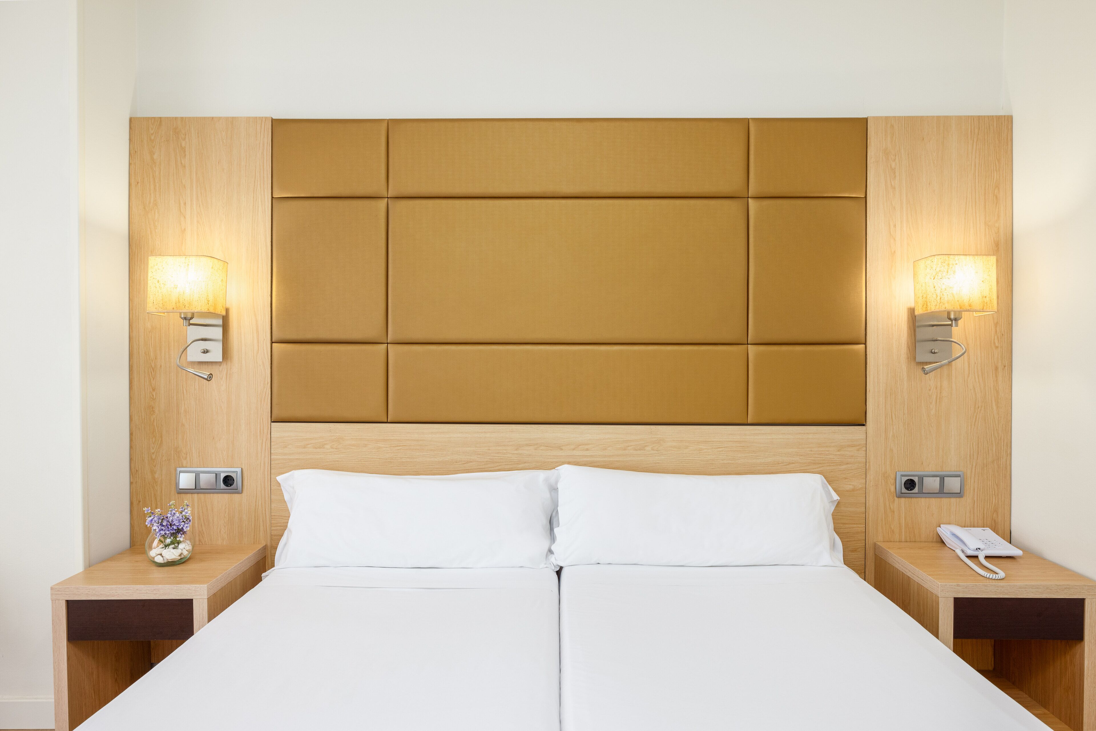 Habitación de hotel con una cama grande, dos mesitas de noche, un cabecero de madera y lámparas fijadas a la pared.