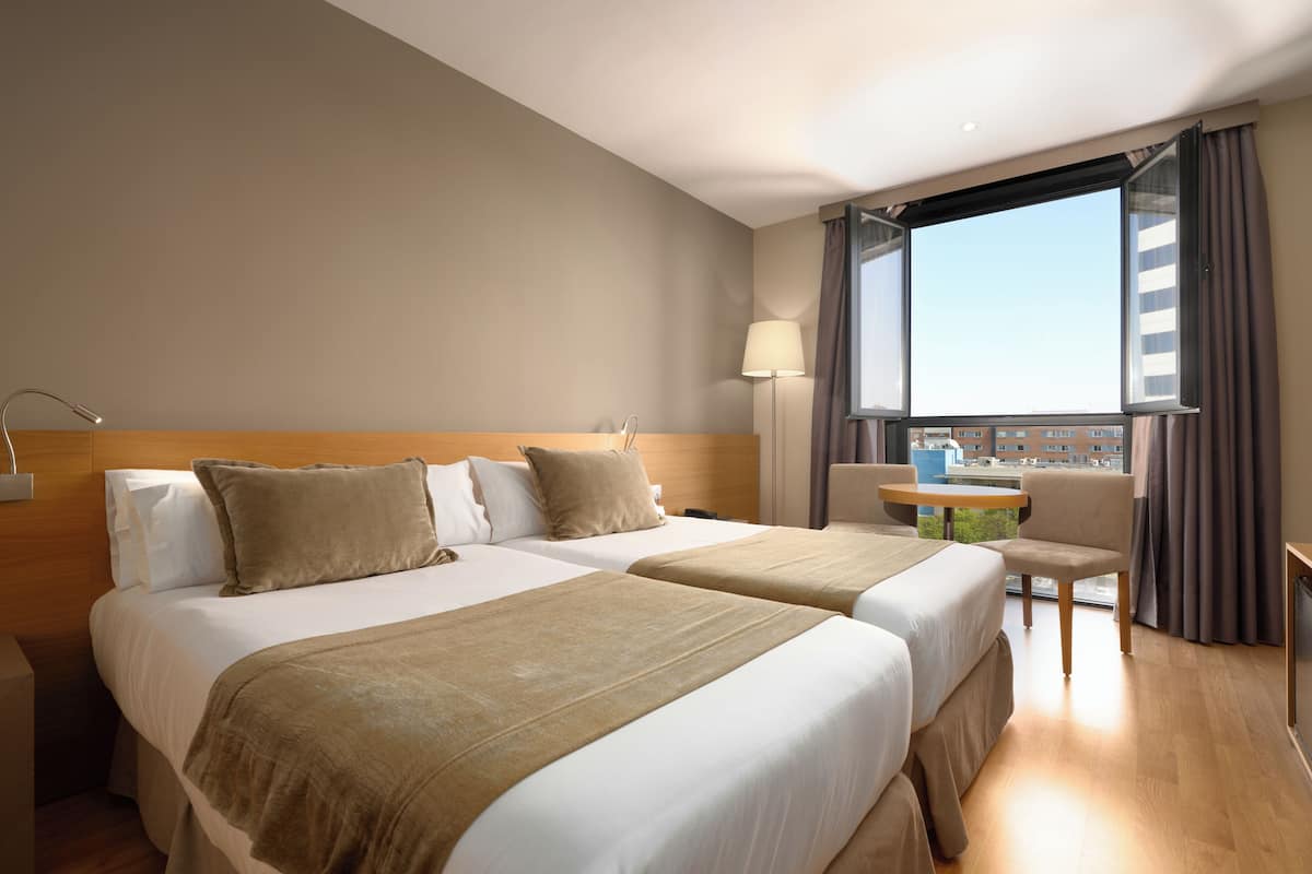 A hotel room with two beds, a desk, a chair, a lamp, and a window with curtains.