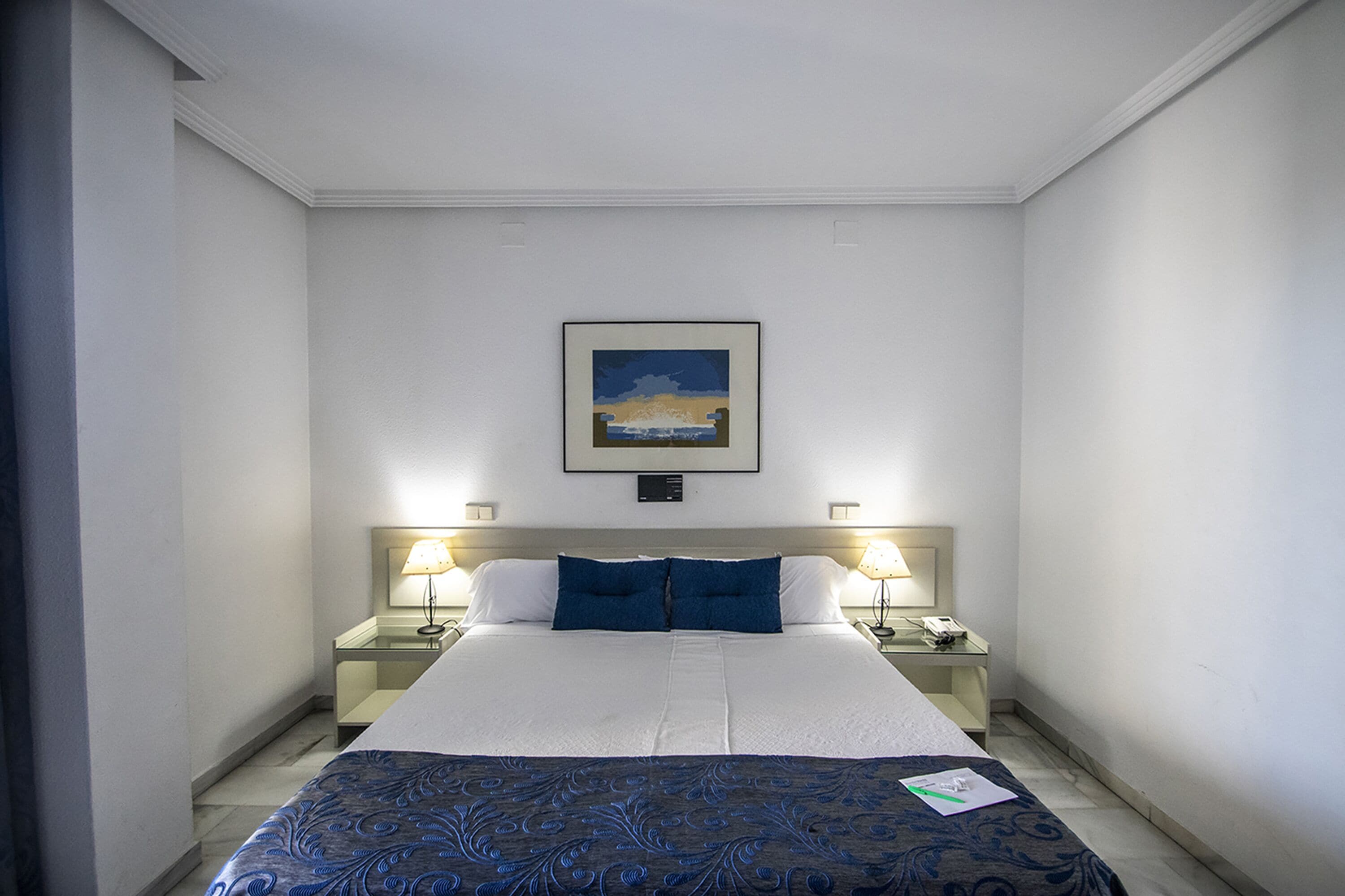 A hotel room with a large bed, two bedside tables, a nightstand, a framed picture on the wall, and a window.