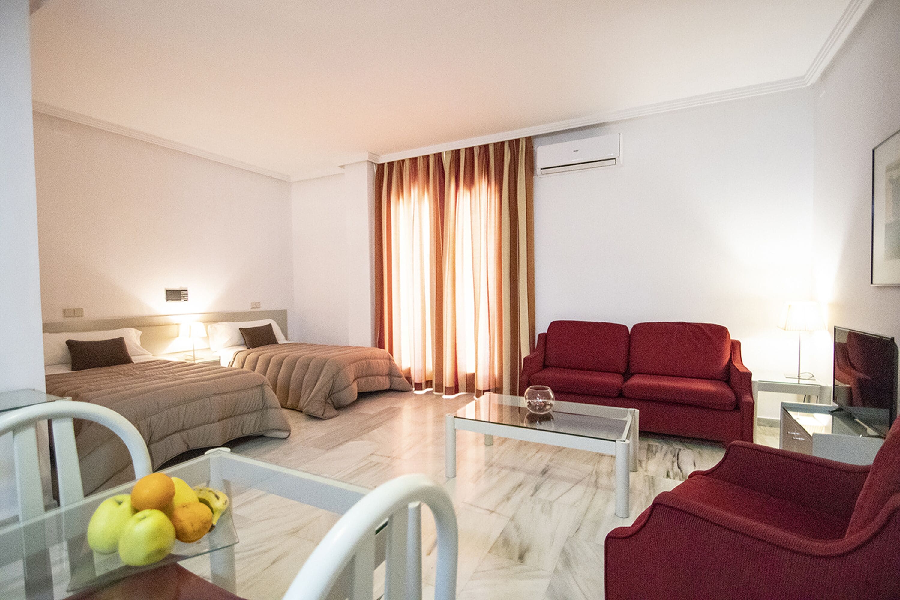 A hotel room with two beds, a red sofa, a glass coffee table, and a fruit bowl.
