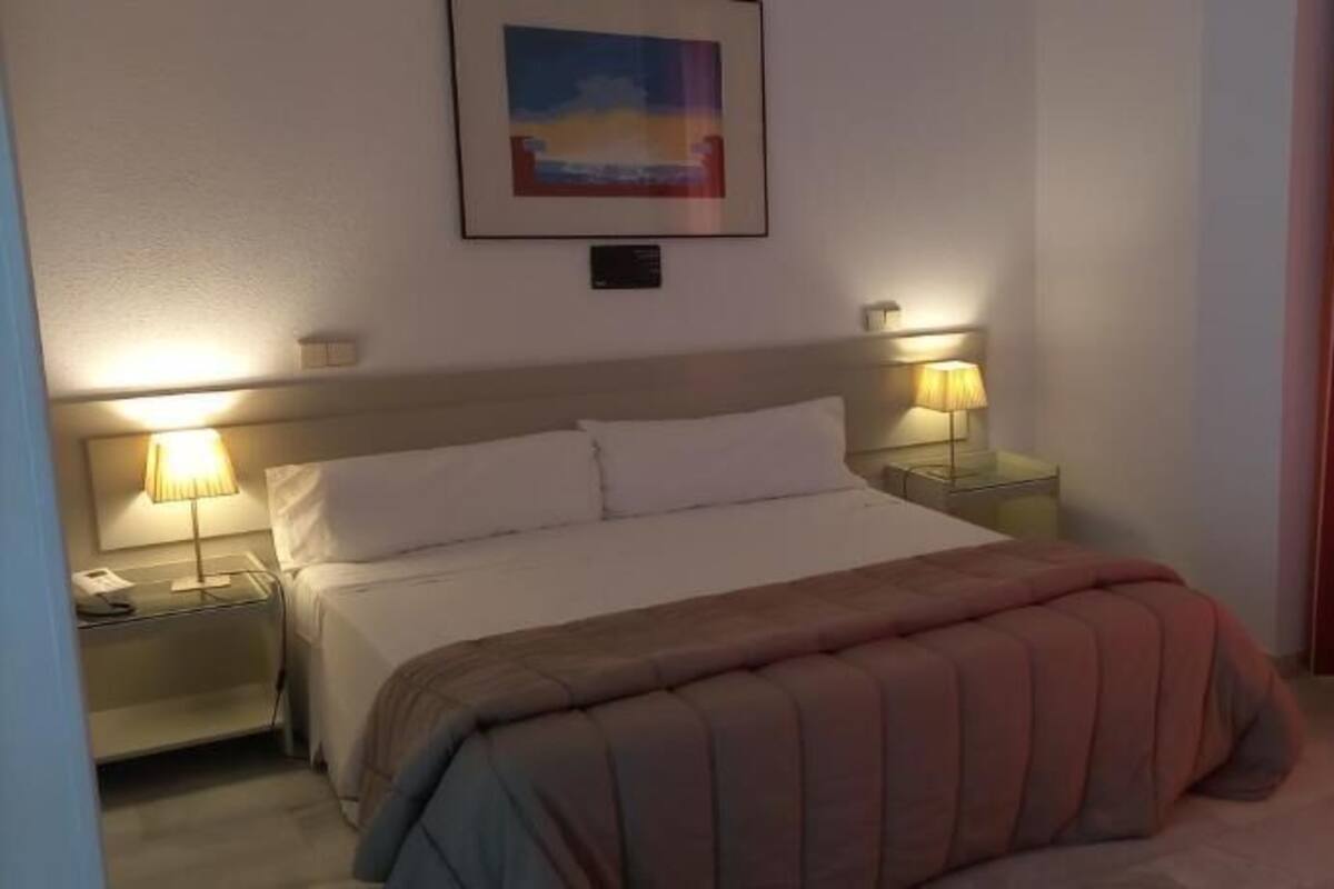 A hotel room with a bed, two bedside tables with lamps, a framed picture on the wall, and a window with curtains.
