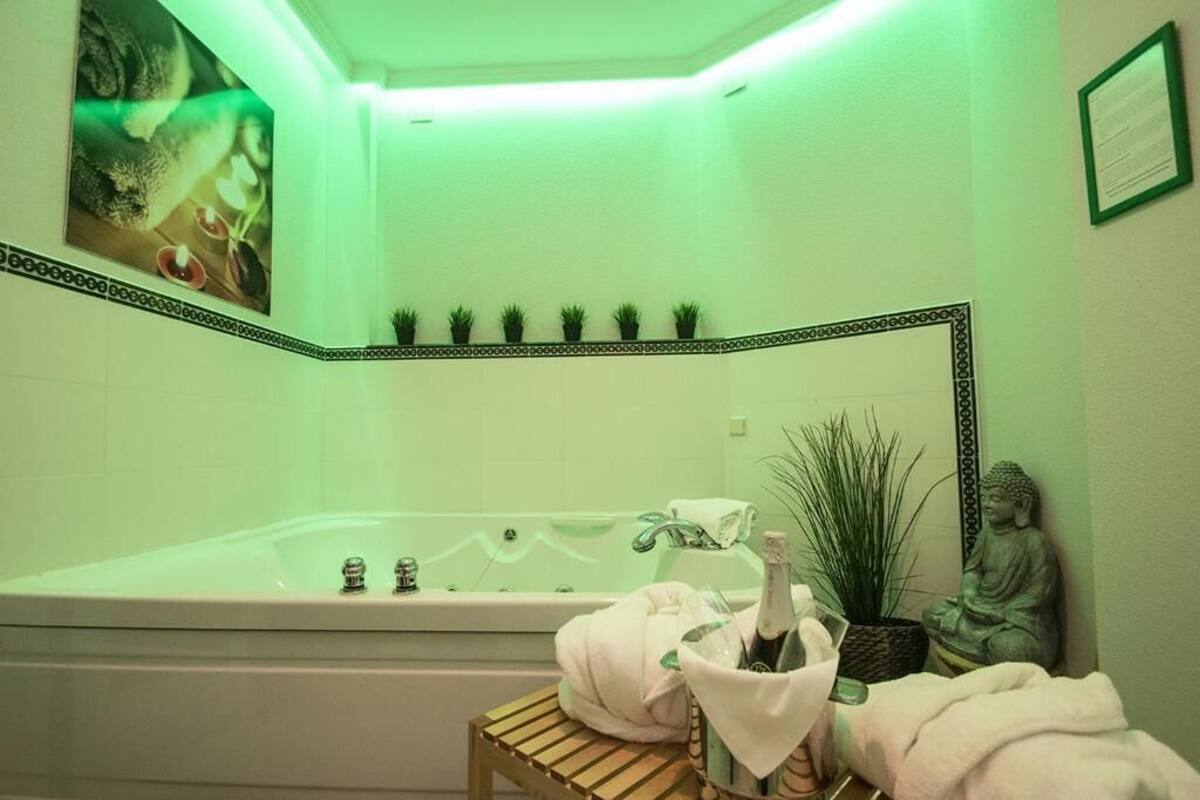 A bathroom with a bathtub, a wooden stool with bottles, a Buddha statue, and a green-lit wall.