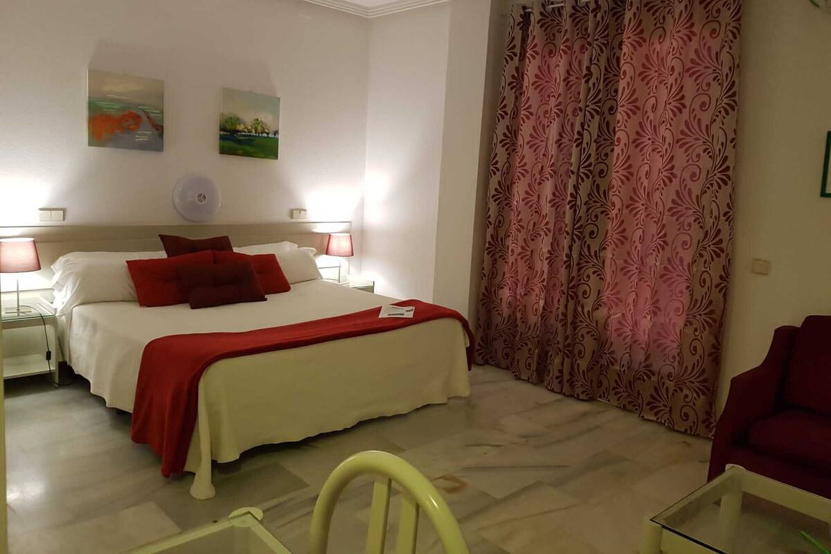 A hotel room with a bed, two bedside lamps, a red chair, a glass table, and a patterned curtain.
