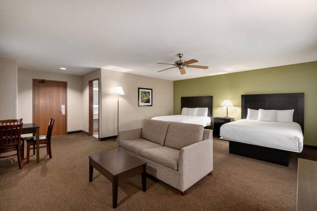 Habitación de hotel con cama, sofá, escritorio, silla, ventilador de techo y mesita de noche con lámpara.