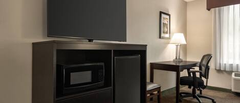 A hotel room with a flat-screen TV, a microwave, a refrigerator, a desk with a chair, a lamp, and a window with curtains.