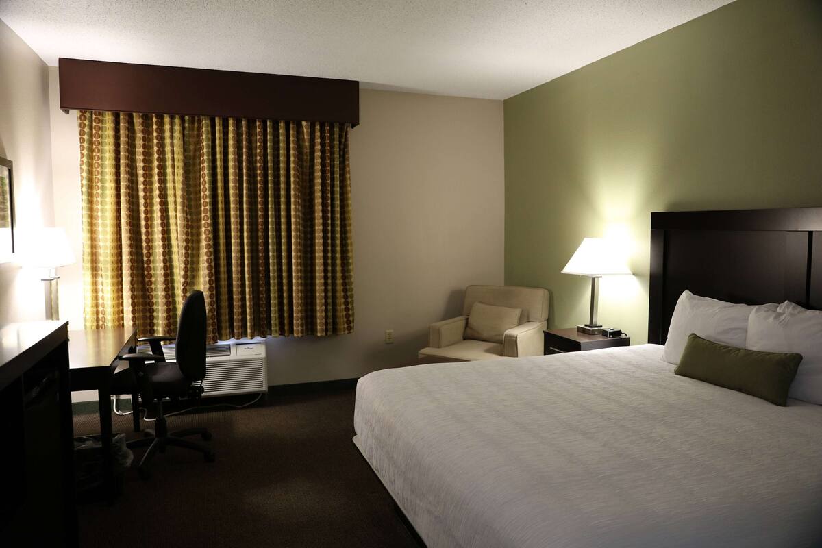 Una habitación de hotel con cama, escritorio, silla, lámpara y ventana con cortinas.