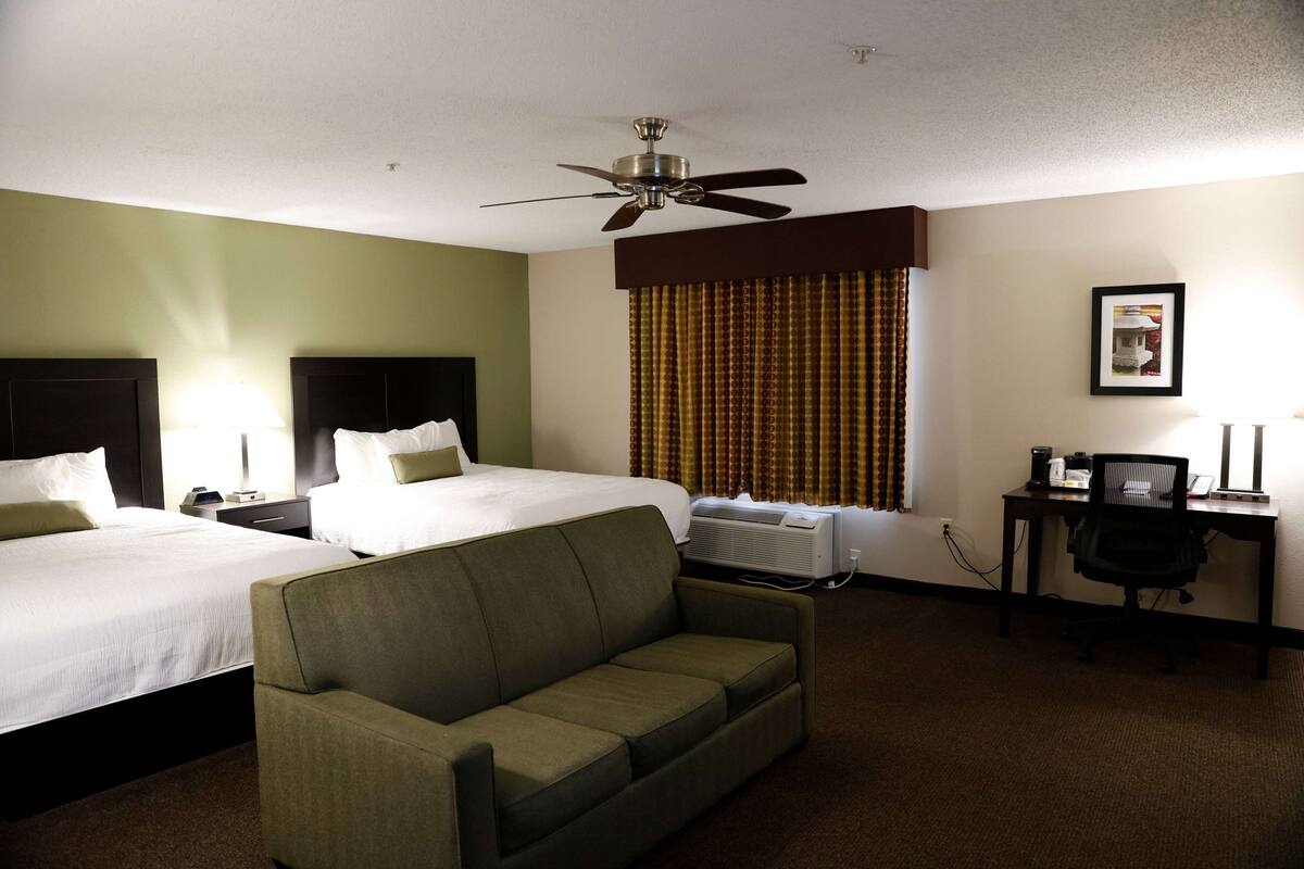Habitación de hotel con dos camas, un sofá, un escritorio con silla, un ventilador de techo y una ventana con cortinas.