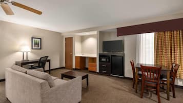A hotel room with a sofa, a dining table with chairs, a kitchenette, a TV, and a ceiling fan.